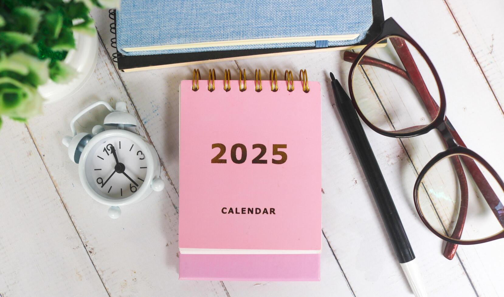 Pink calendar 2025 on office desk with notebook, glasses, pen and alarm clock. New year 2025 with office desk concept photo
