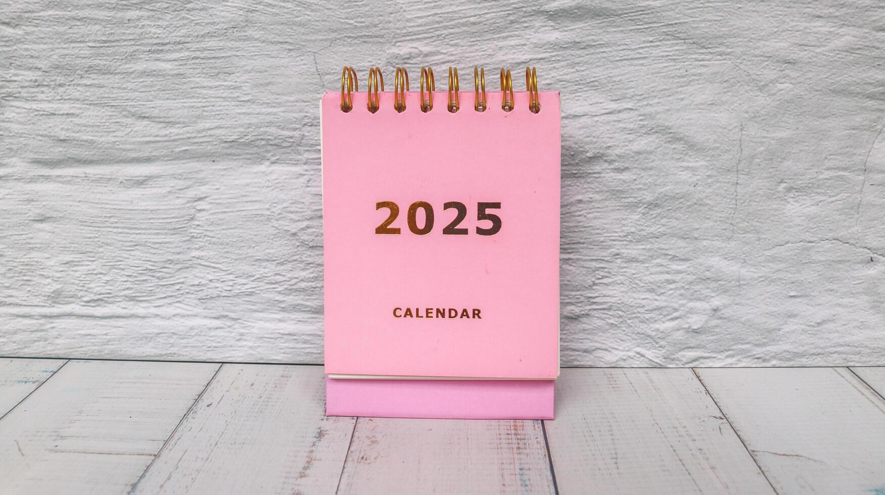 Pink calendar 2025 standing on office desk with flower pot and alarm clock. New year 2025 with office desk concept with table background. photo
