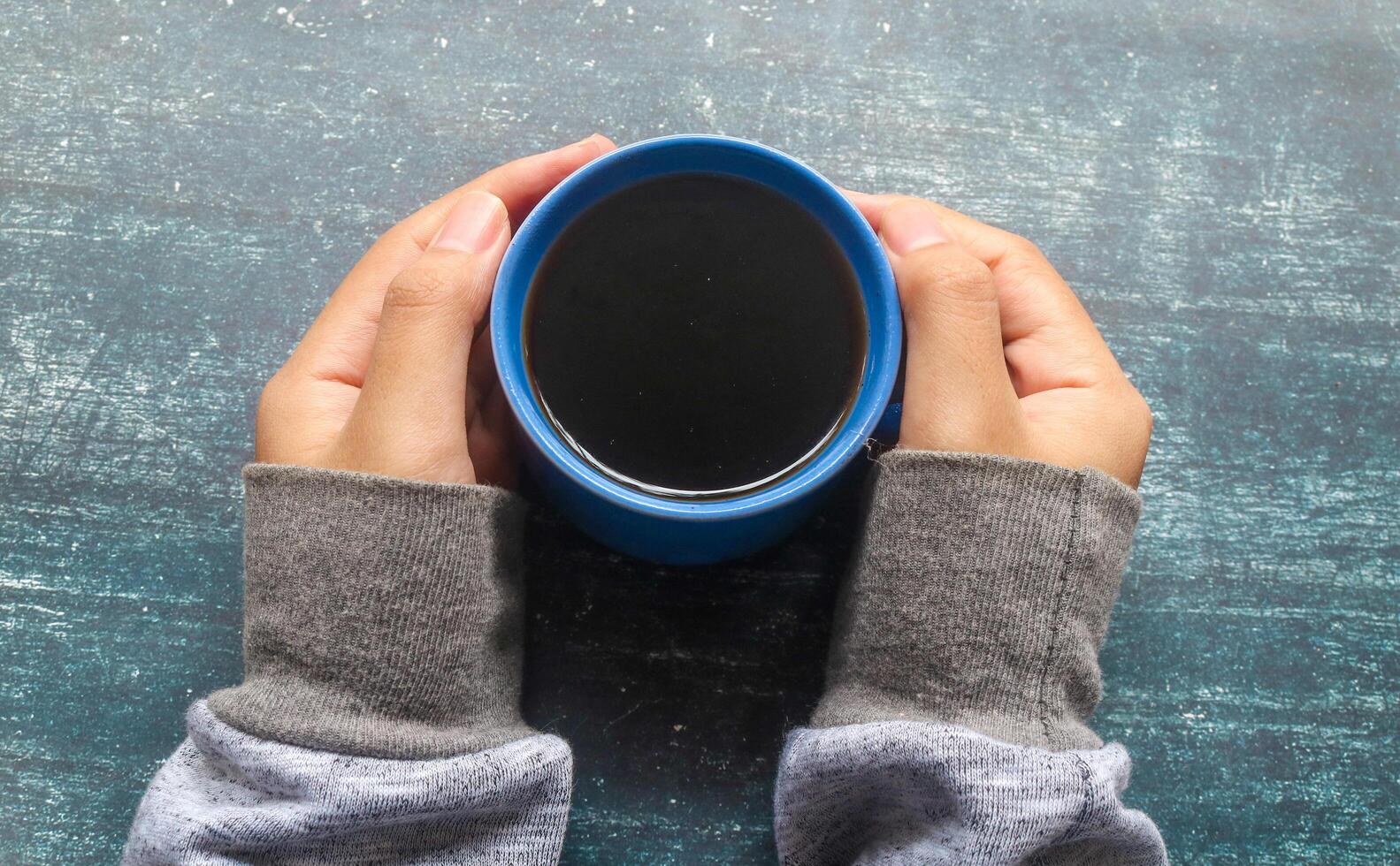 Hand holding a cup of black coffee in a blue cup with a gray background. Holding a cup of black coffee in the morning when the weather is cold photo