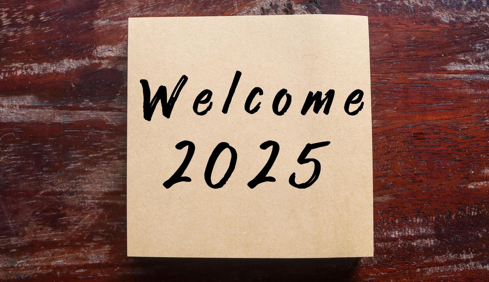 Brown sticky note with Welcome 2025 written on wooden table. Welcoming new year 2025 concept photo
