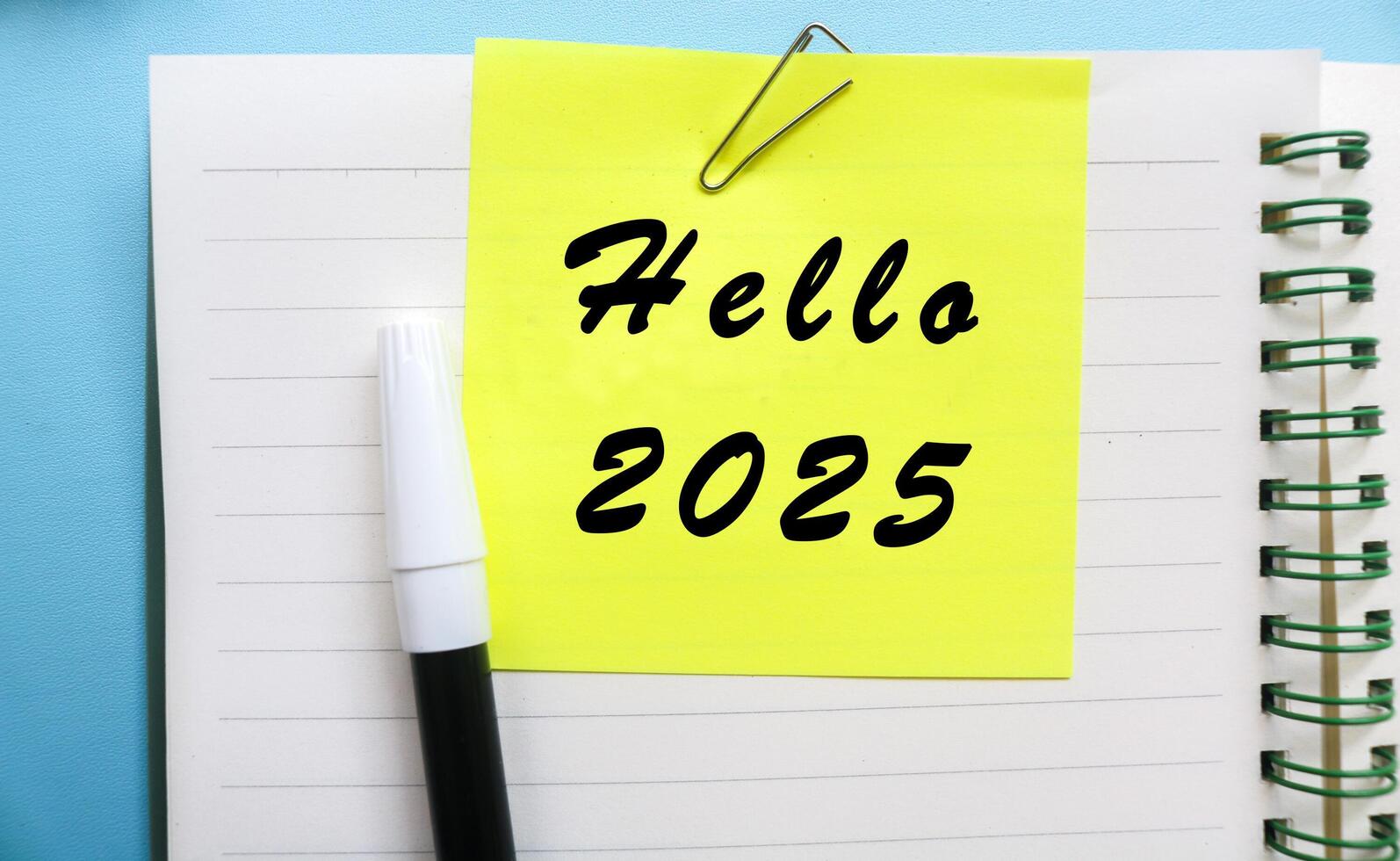 Yellow sticky note with Hello 2025 written on a notebook with stationery isolated on blue background. Welcoming the new year 2025 concept photo