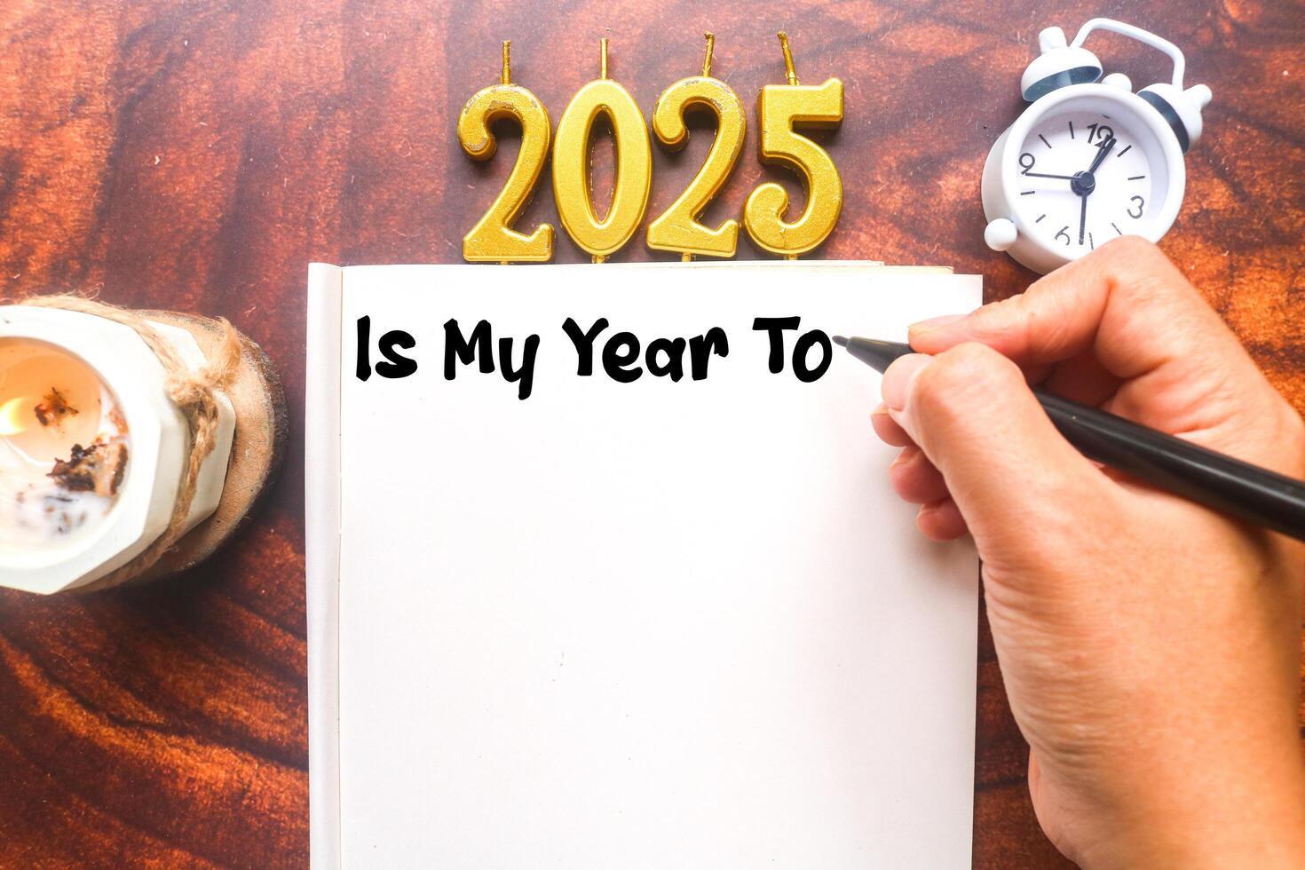 A person writes 2025 Is my year to in a notebook surrounded by aromatherapy candles and clocks. Planning to do list for 2025, life goals for 2025 photo