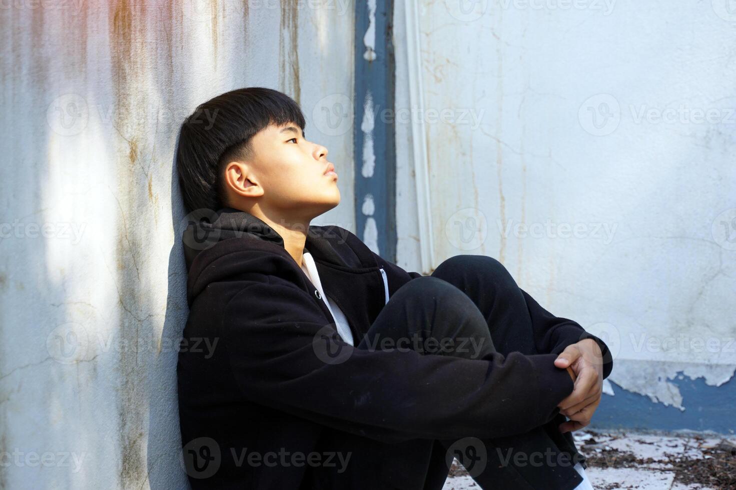 teenage boy sits, hugging his knees with his back against the wall of an abandoned building, alone. Concept of lonely, abandoned, quarantined, depressed. photo