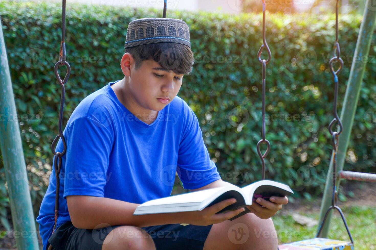 A young Muslim boy wears a blue T-shirt and a gray Taqiyah cap. Sitting and posing for photos while reading a book at the park.