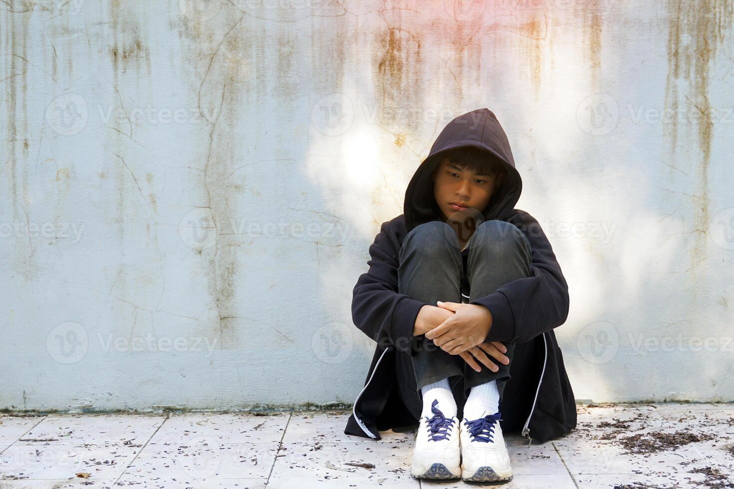 teenage boy sits, hugging his knees with his back against the wall of an abandoned building, alone. Concept of lonely, abandoned, quarantined, depressed. photo