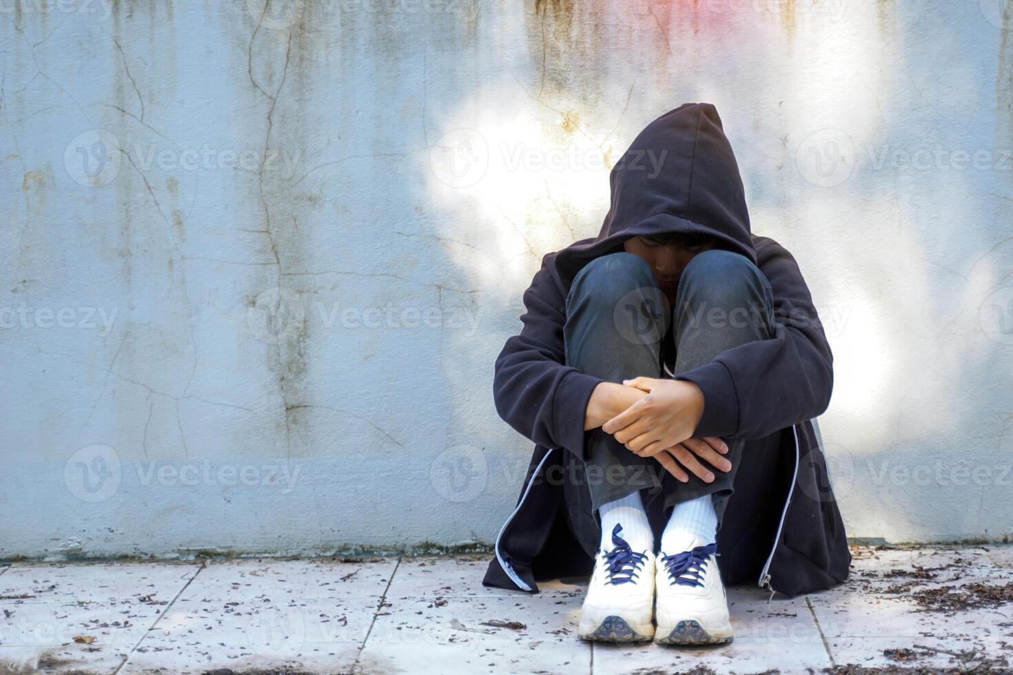 teenage boy sits, hugging his knees with his back against the wall of an abandoned building, alone. Concept of lonely, abandoned, quarantined, depressed. photo