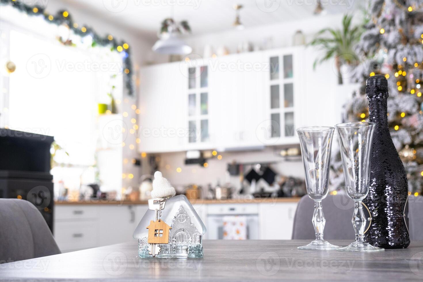 Ice transparent house with a key on table of cozy white kitchen with Christmas tree and Christmas decor with garland lights. Housing, interior, renovation, mortgage, rent in New Year photo