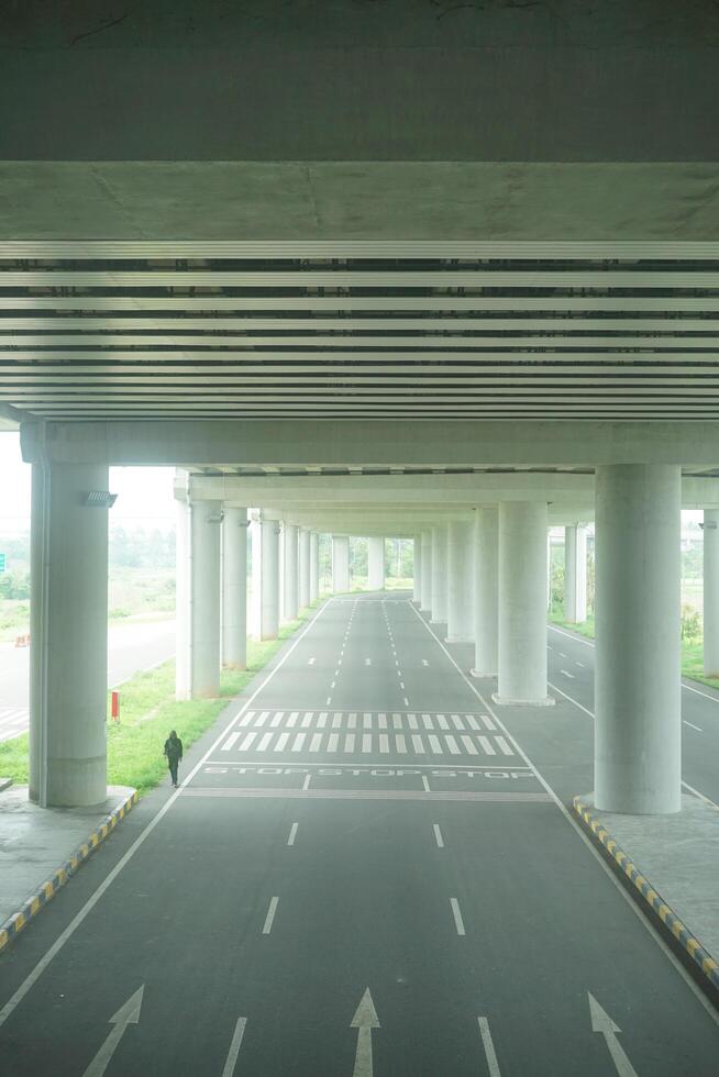 A long, straight road passing through a concrete overpass structure with multiple arched openings, surrounded by green foliage at new yogyakarta international airport photo