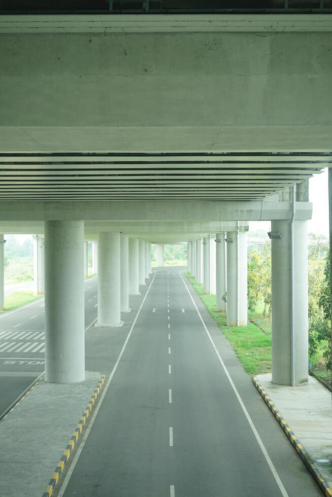 A long, straight road passing through a concrete overpass structure with multiple arched openings, surrounded by green foliage at new yogyakarta international airport photo