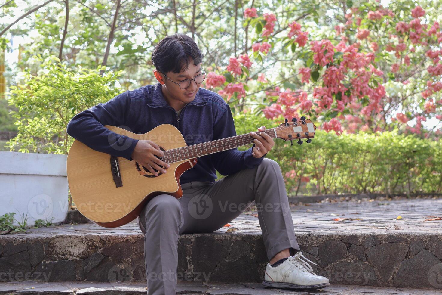 Asian man plays his guitar in a city park on a warm summer day, surrounded by greenery. The serene atmosphere, bright sunlight, and soothing melody create a peaceful and vibrant moment photo