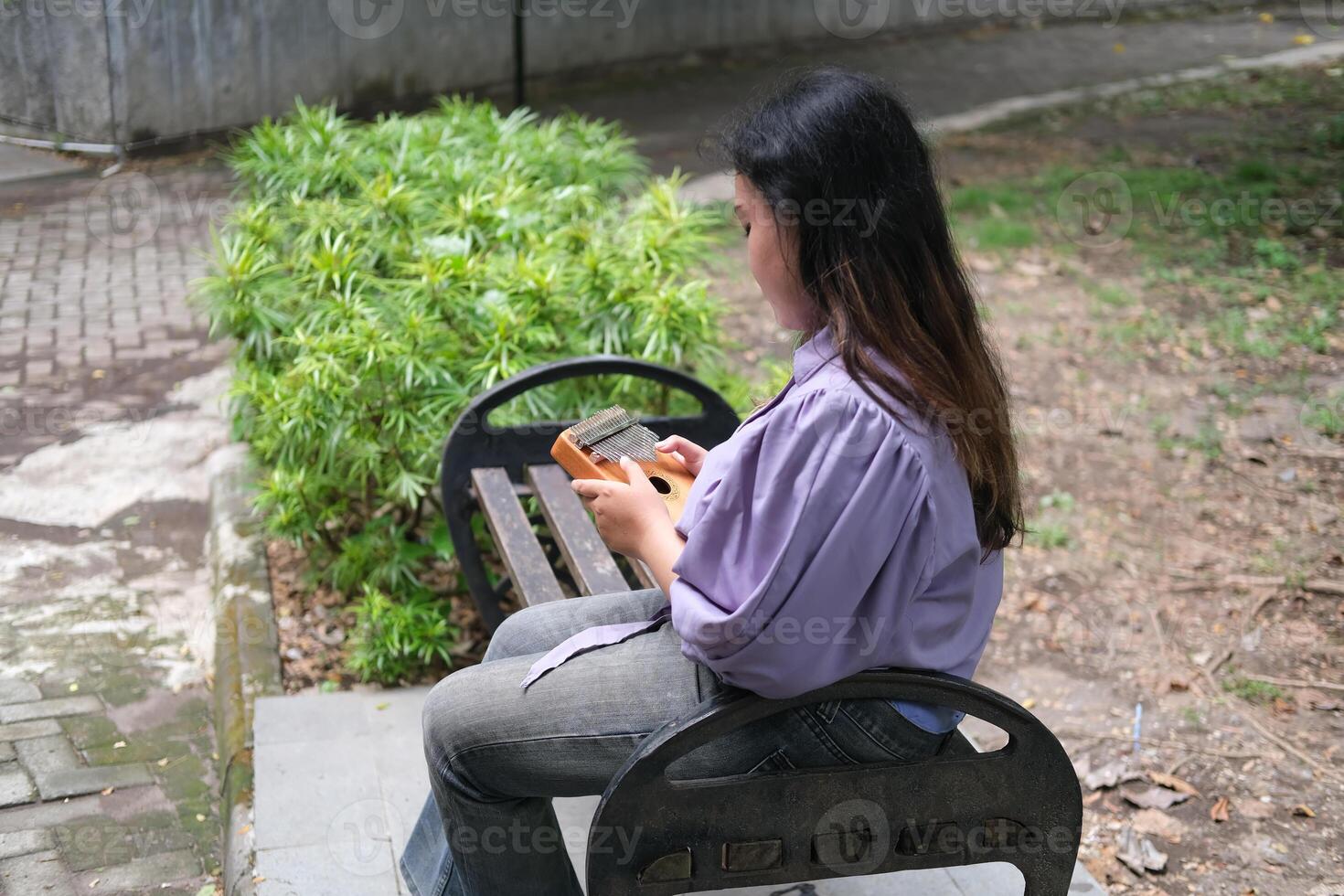 young Asian woman plays a kalimba while relaxing in a green city park during summer. The bright sunlight, lush surroundings, and soft melodies create a calm and refreshing outdoor scene photo