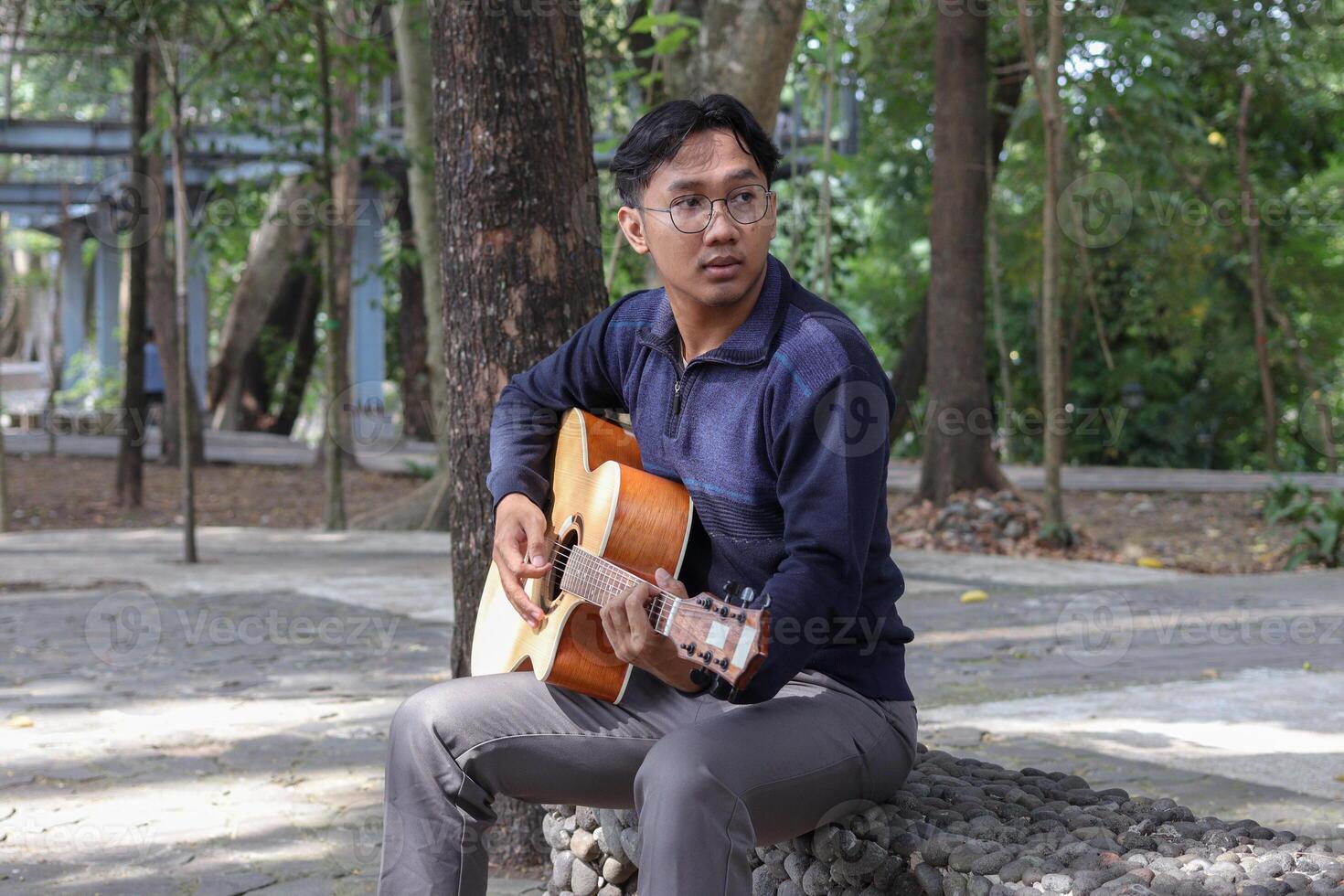 Portrait of Asian man playing guitar in a park during summer, surrounded by lush greenery and the peaceful atmosphere of the season photo