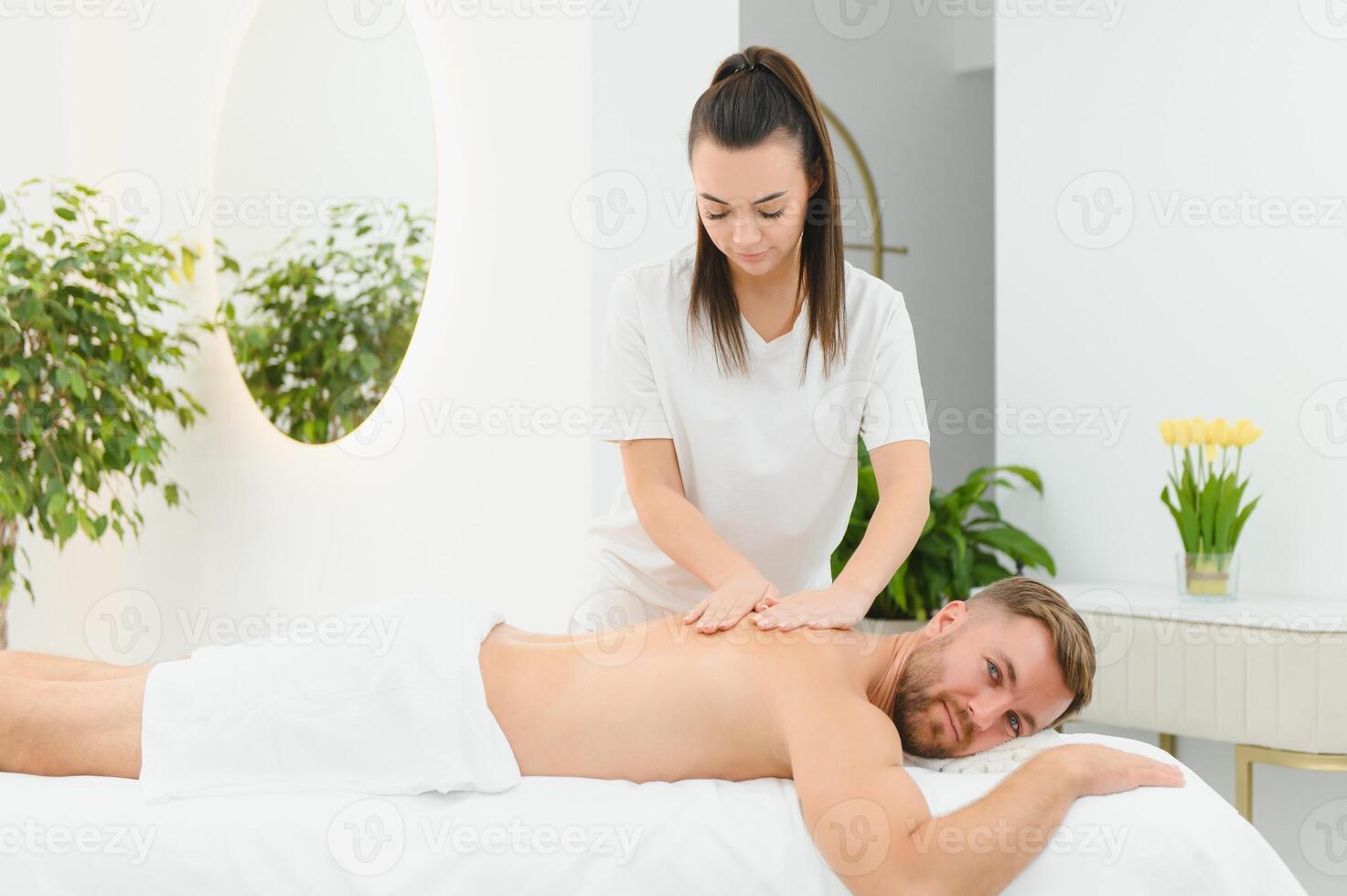 Female masseur with long hair in white uniform doing back massage to man lying on couch in bright salon room at daytime photo