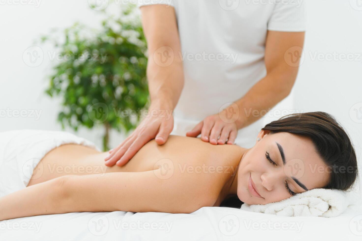 Woman enjoying professional body massage at a modern spa salon. Relaxed young lady lying on a massage table while a skilled masseur is gently massaging her back. Beauty, health, pleasure, concept photo