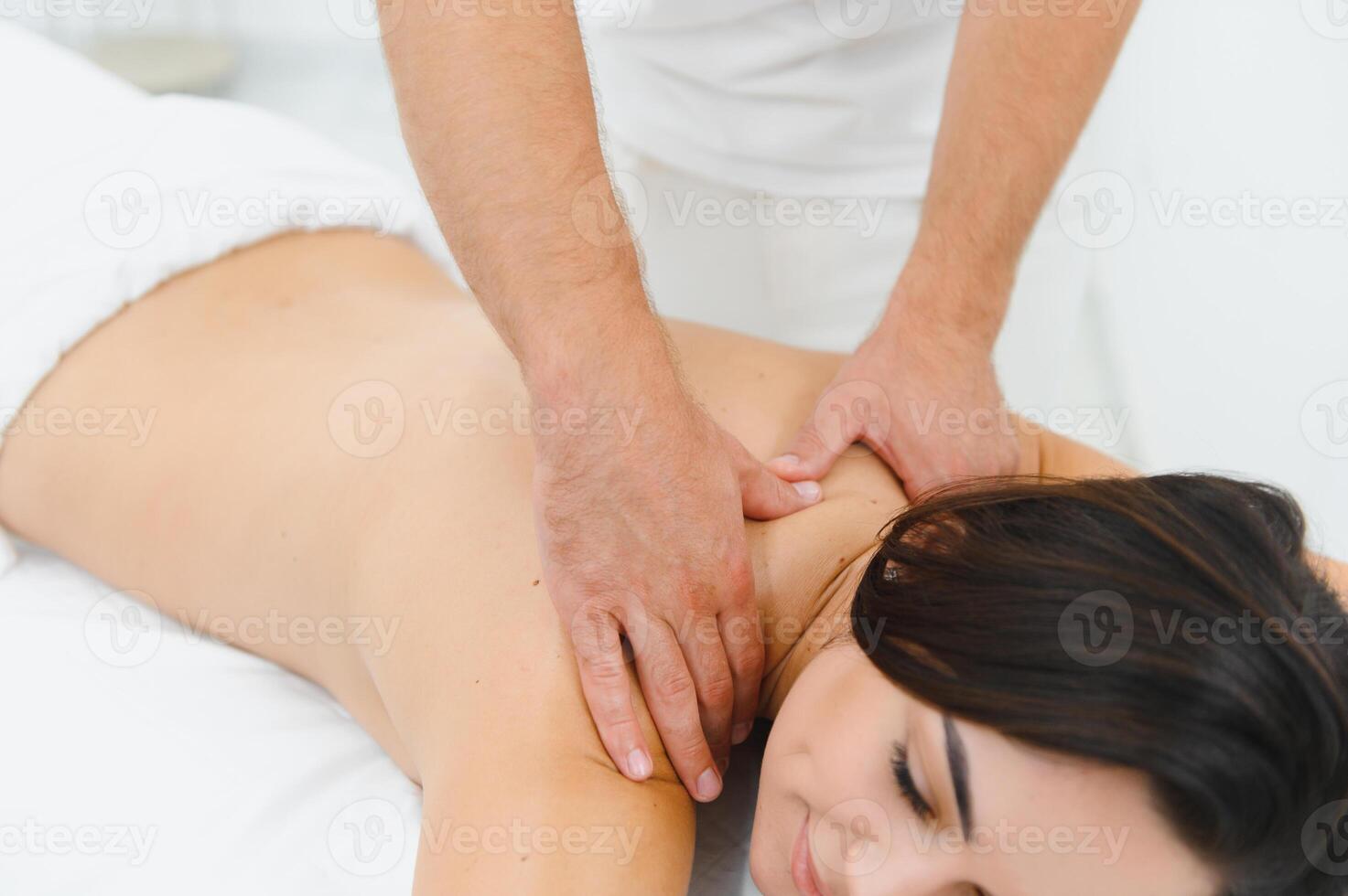 Woman enjoying professional body massage at a modern spa salon. Relaxed young lady lying on a massage table while a skilled masseur is gently massaging her back. Beauty, health, pleasure, concept photo