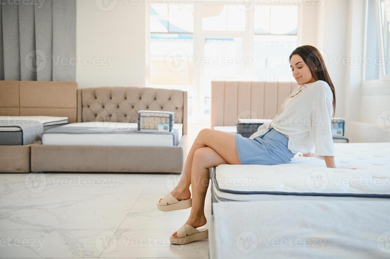 happy brunette female is sitting on mattress for testing it in the store photo