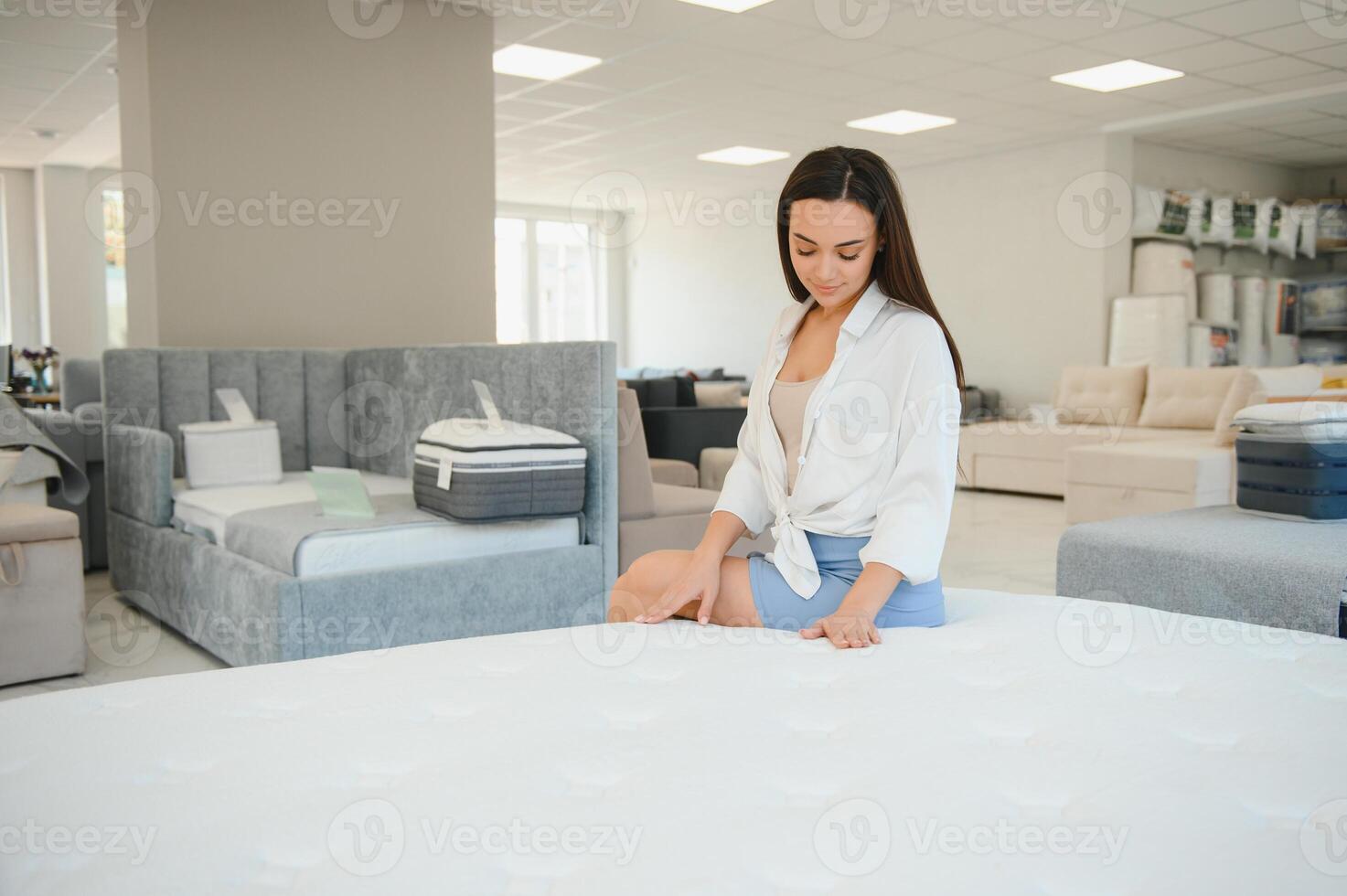 happy brunette female is sitting on mattress for testing it in the store photo