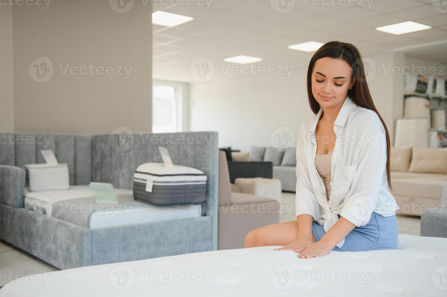 happy brunette female is sitting on mattress for testing it in the store photo