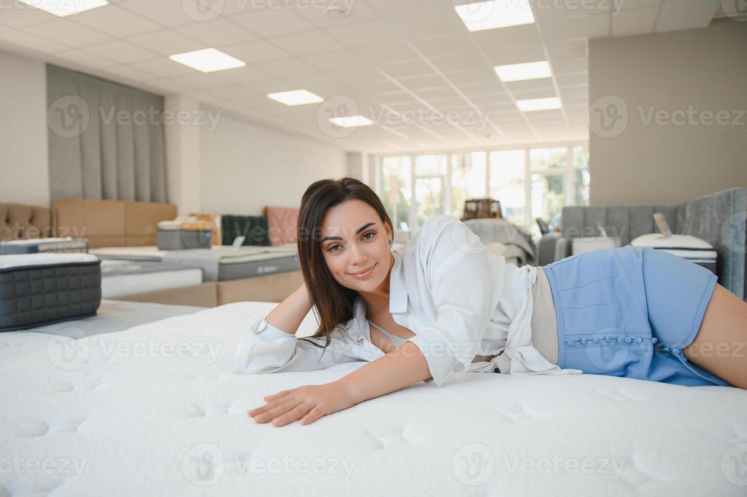 happy brunette female is sitting on mattress for testing it in the store photo