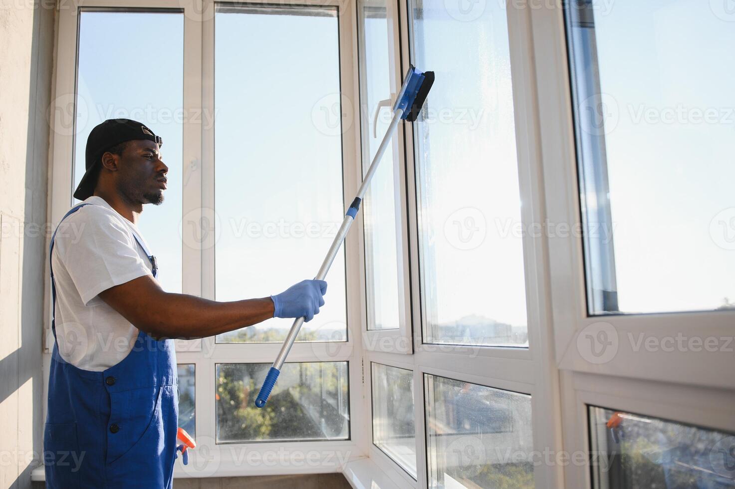 African Male professional cleaning service worker in overalls cleans the windows and shop windows of a store with special equipment photo