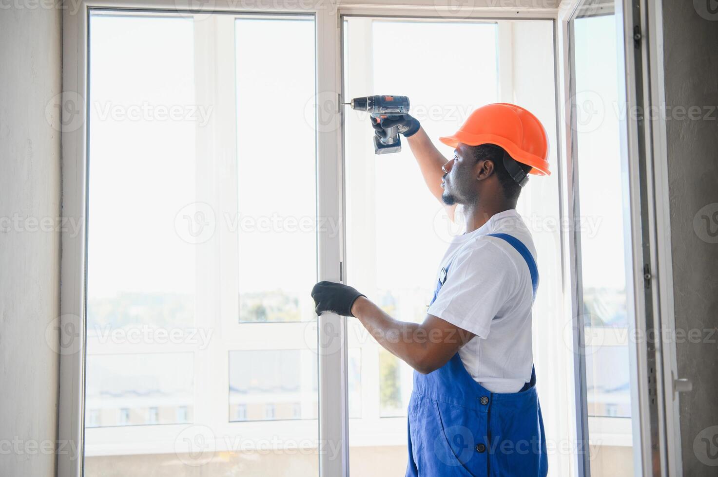 African Workman in overalls installing or adjusting plastic windows in the living room at home photo