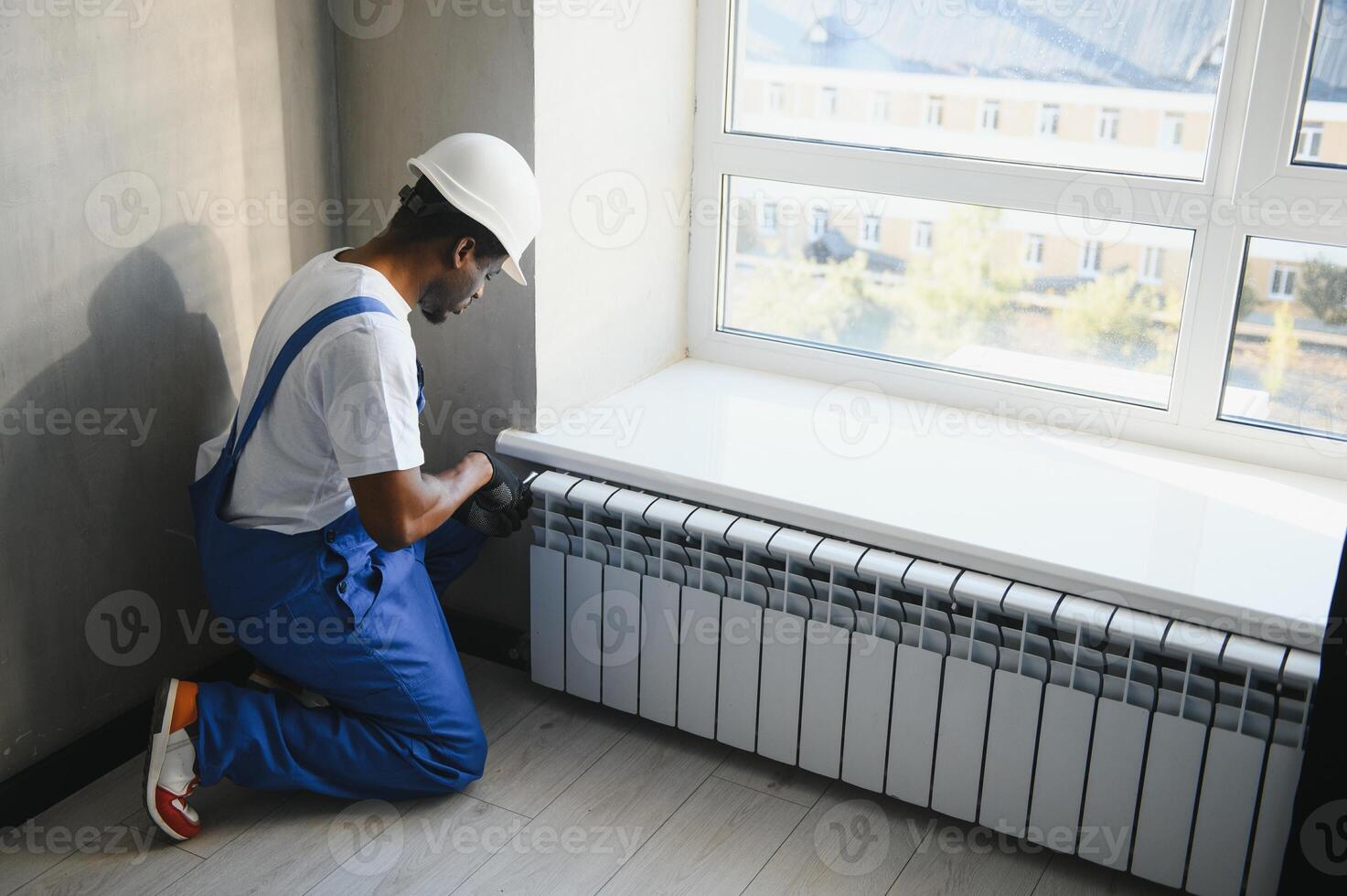 afro repairman in blue using tools while installing or repairing heating radiator. plumber installing heating system in apartment. photo
