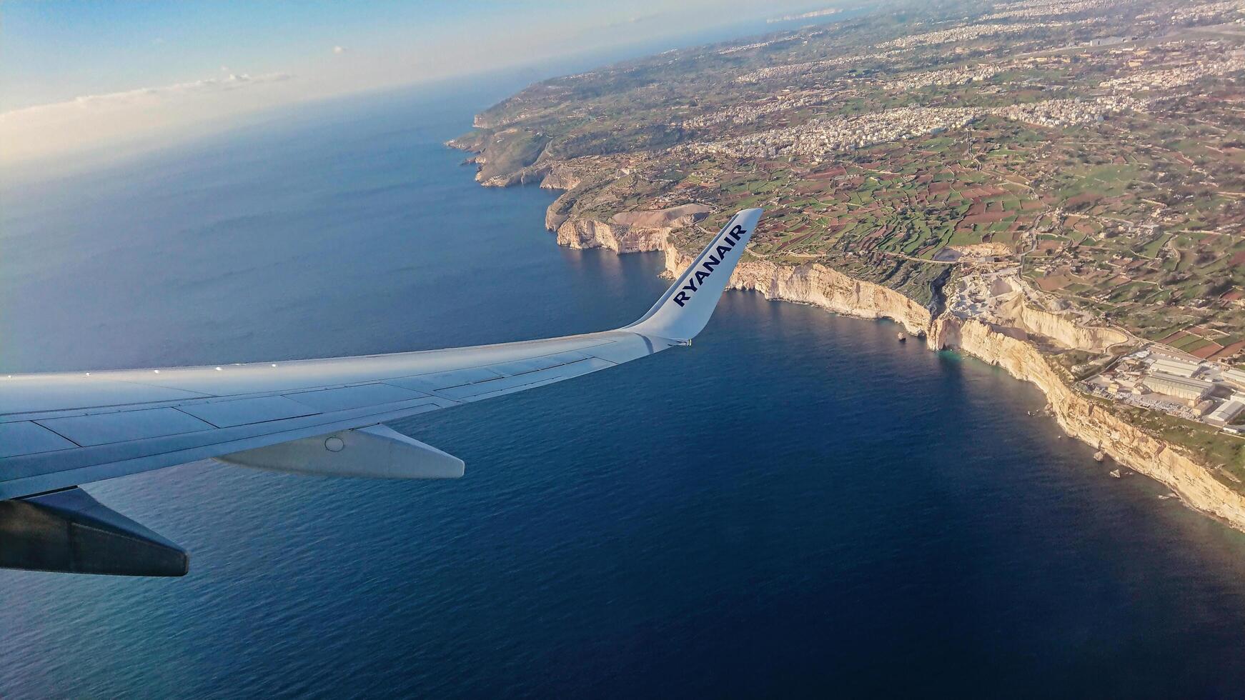 Luqa, Malta 9.01.2020. Ryanair airplane wing soaring over picturesque Maltese coastline cliff, showcasing deep blue sea and clear sky during flight photo