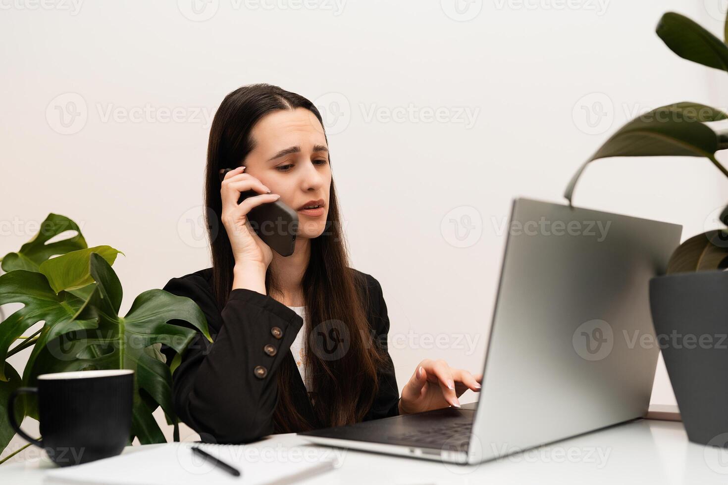 Upset woman speaking on the phone while working on computer laptop in office with white walls and green plant photo