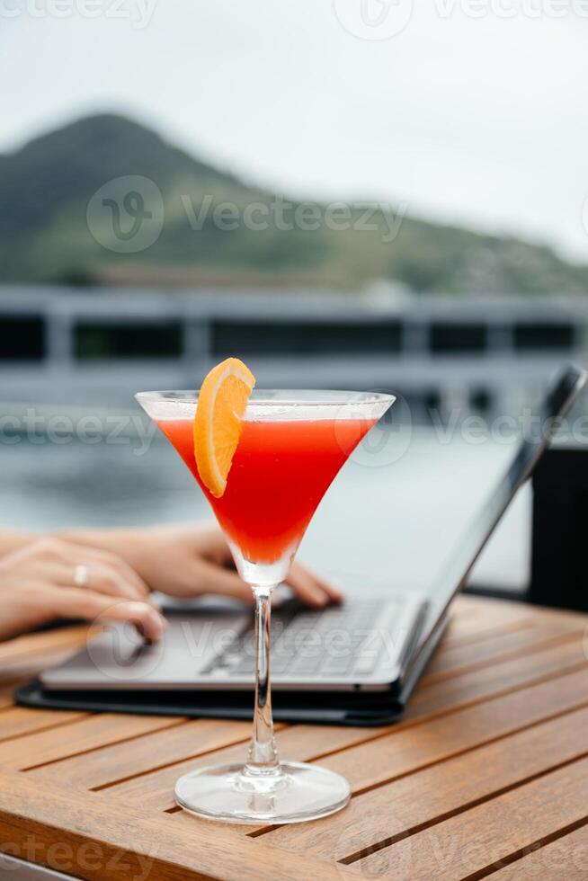 Female hands works with Laptop and glass of refreshing red cocktail on a table background. Work and travel or remote business concept. Out of office work concept. Vertical shot photo