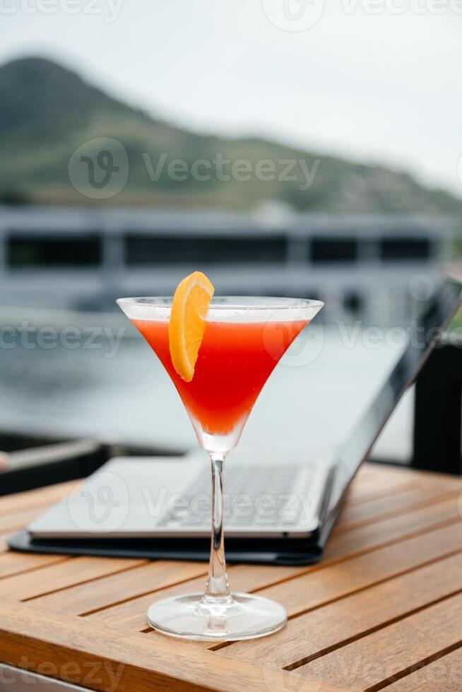 Laptop and glass of refreshing red cocktail on a table background. Work and travel or remote business concept. Out of office work concept. Vertical shot photo