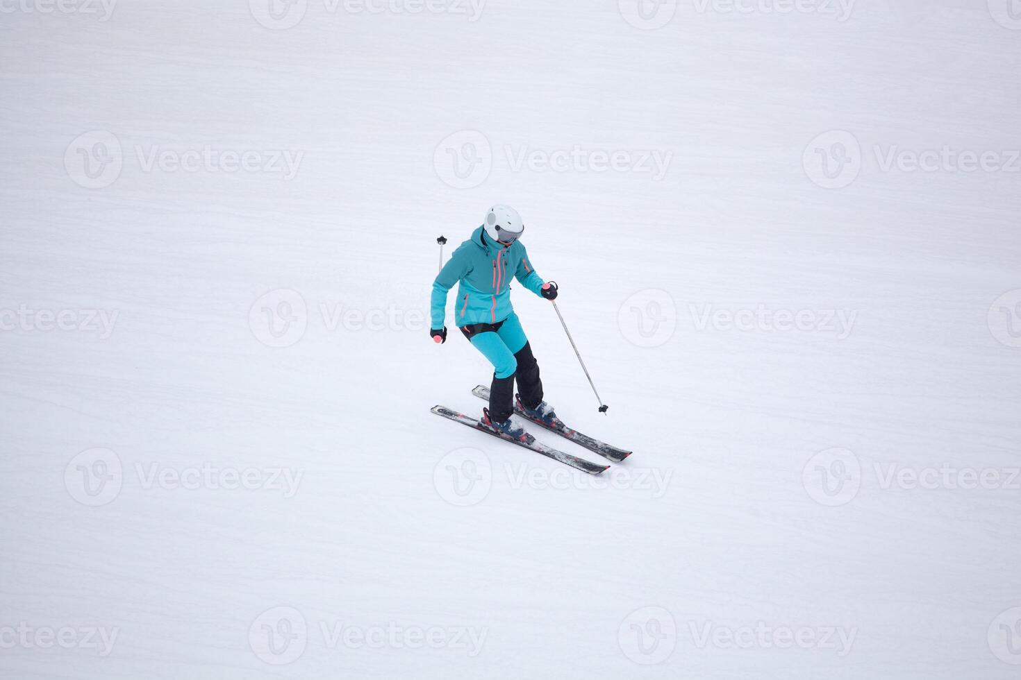 A skier in a turquoise outfit and white helmet skiing down a snowy slope, holding ski poles photo