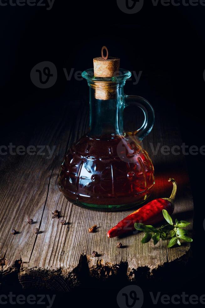 A rustic glass bottle filled with dark liquid, sealed with a cork, sits on a wooden surface next to a red chili pepper and some herbs. photo