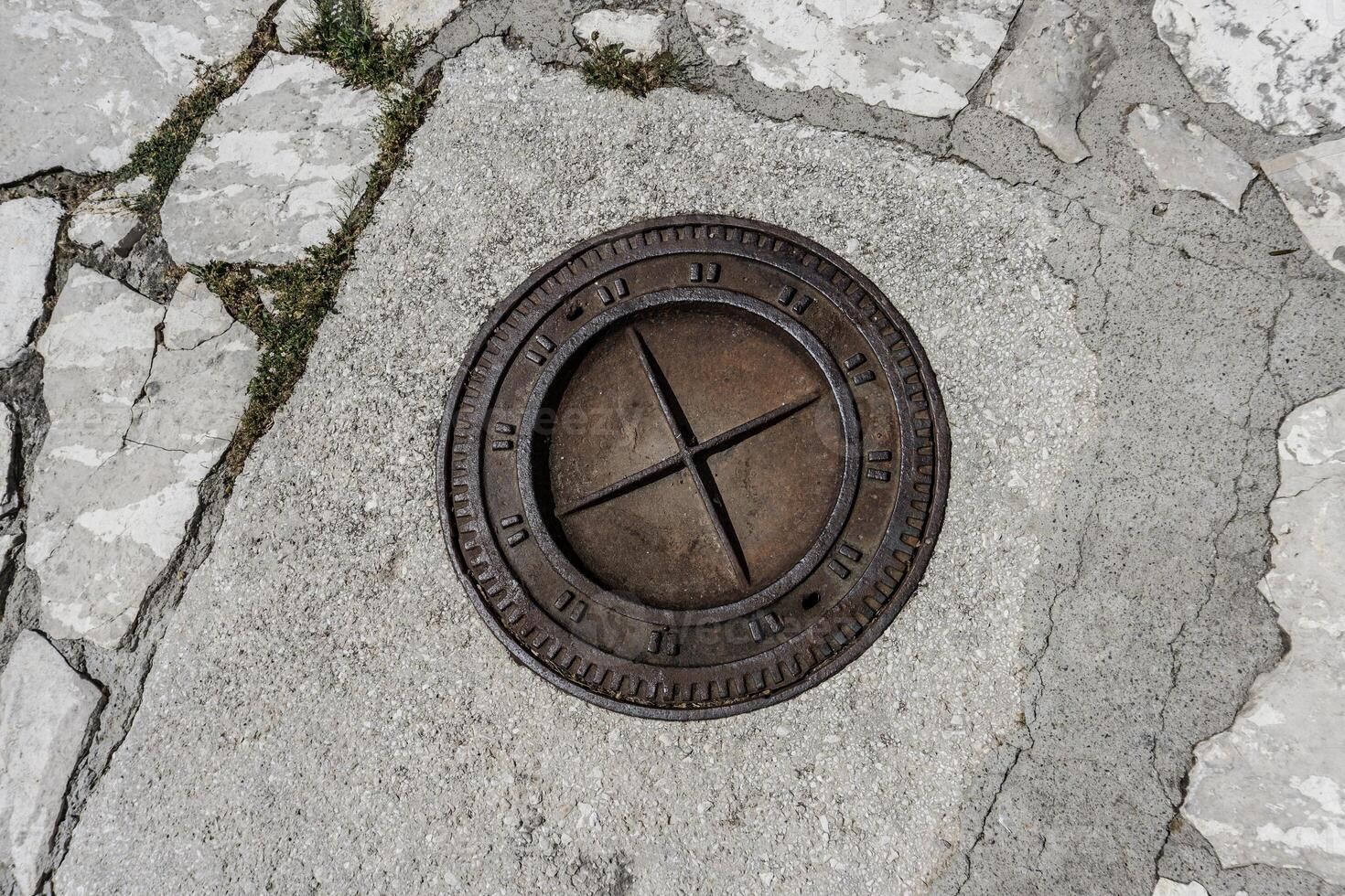 A round metal manhole cover with a cross design in the center, embedded in a cracked concrete surface with some grass growing between the cracks photo