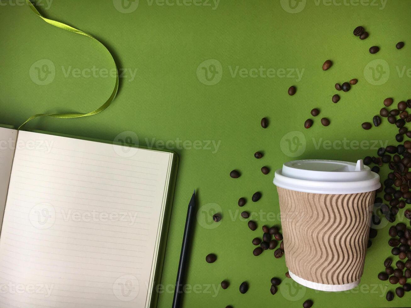 An open lined notebook with a pencil, takeaway coffee cup with a wavy pattern, and scattered coffee beans on a green background. photo