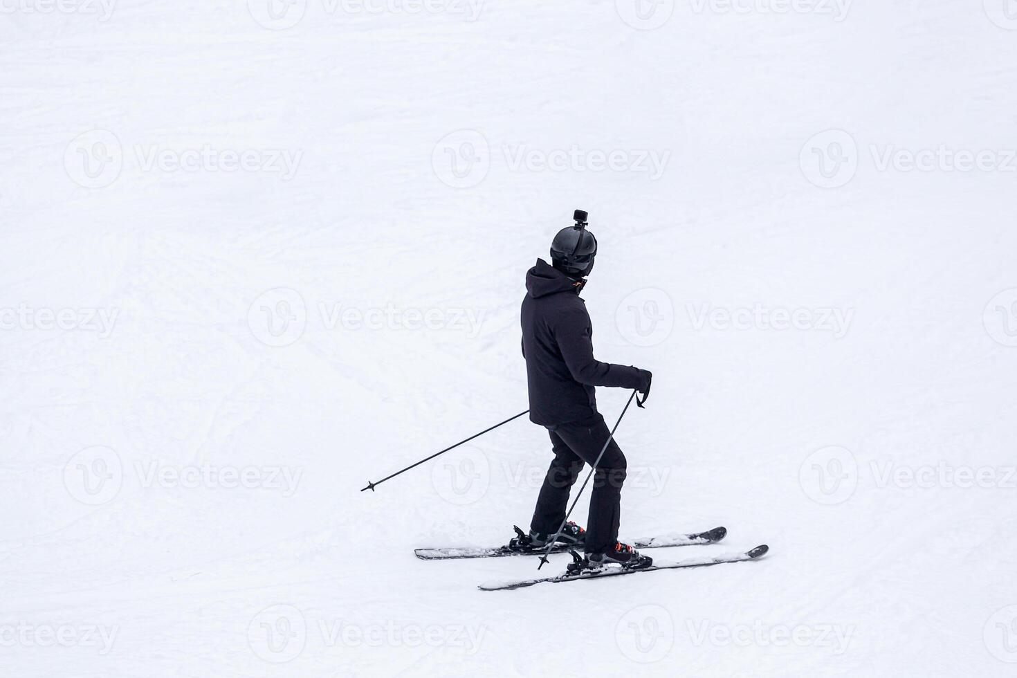 A skier dressed in black gear, including a helmet with a mounted camera, is skiing on a snowy slope, creating a dynamic and adventurous winter sports scene photo