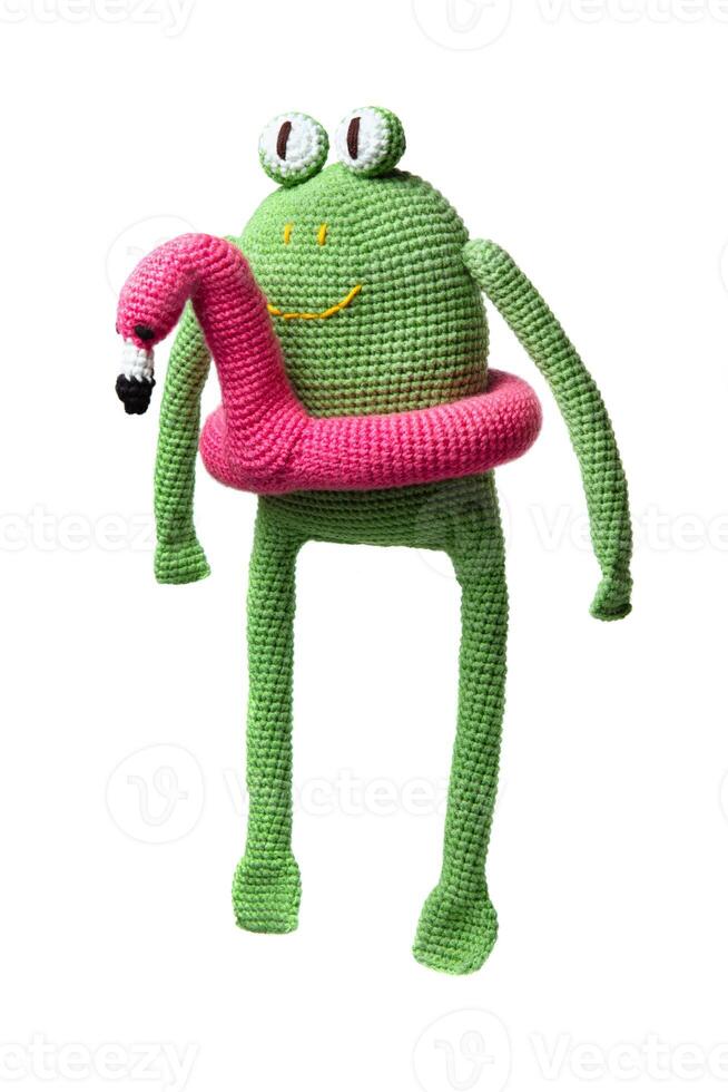 A crocheted green frog toy with large eyes and a pink flamingo float around its body, isolated on a white background. photo