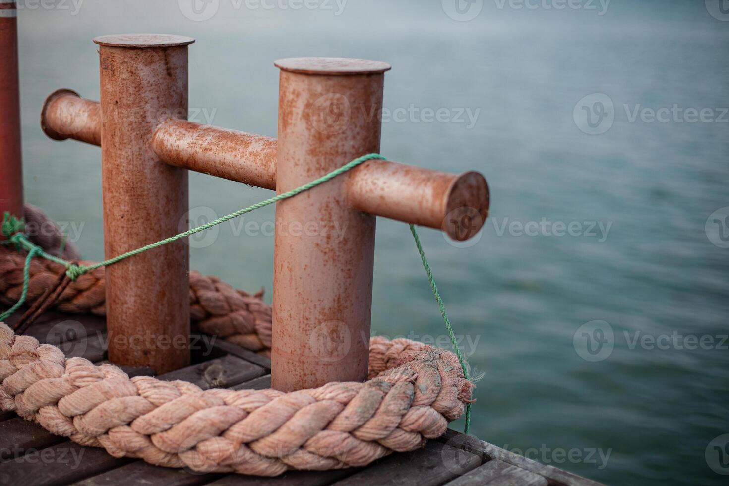 Rusty bollards on a dock with thick ropes and green string, close-up of weathered metal, showcasing nautical and maritime equipment in a harbor setting. photo