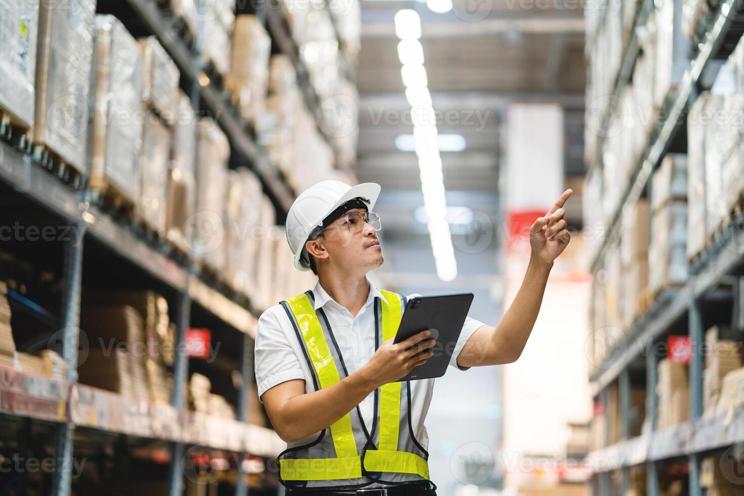 A man in a yellow vest is pointing at a box on a shelf. He is wearing a hard hat and safety glasses photo