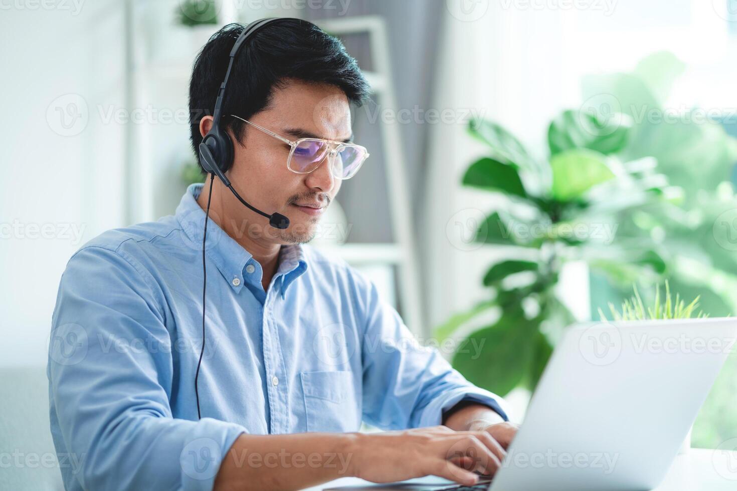 A man wearing a headset and glasses is typing on a laptop. He is focused on his work and he is in a professional setting photo