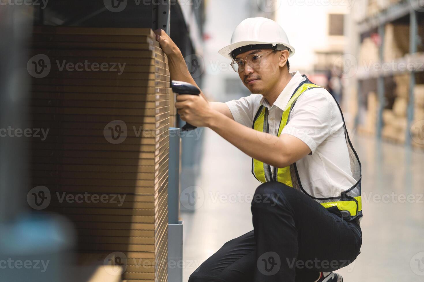A man in a safety vest is sitting on the floor in a warehouse. He is holding a barcode scanner and looking at a box photo