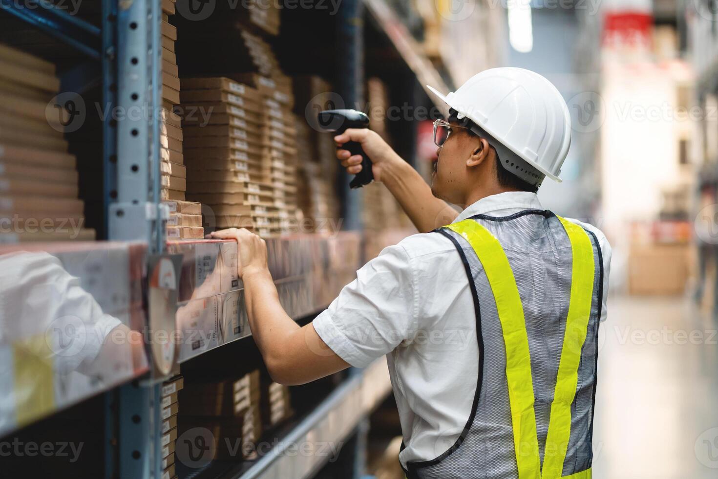 A man in a white shirt and yellow vest is scanning a barcode with a handheld device. He is wearing a hard hat and safety vest, indicating that he is working in a warehouse or industrial setting photo