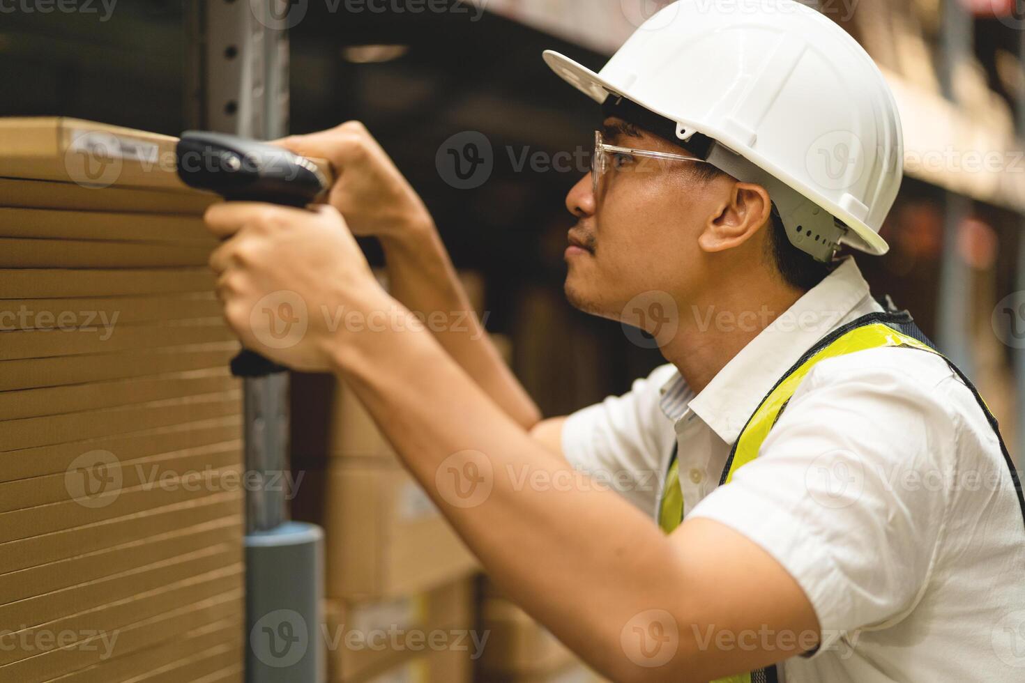 A man in a white shirt and a yellow vest is using a barcode scanner. He is wearing a hard hat and safety glasses photo
