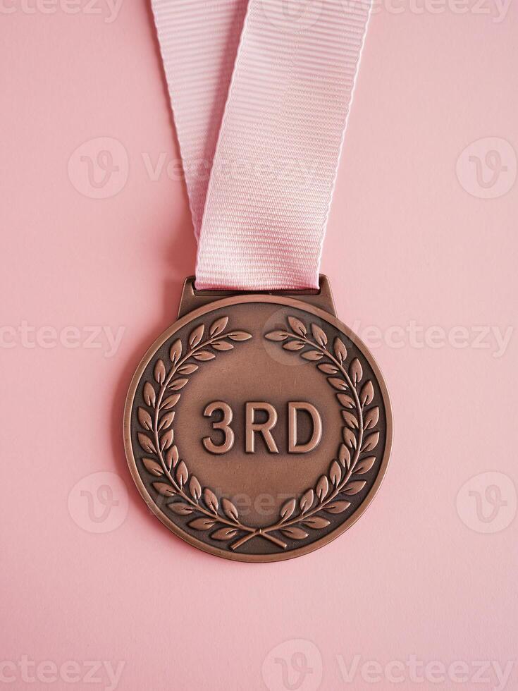 Bronze medal with '3RD' engraved, hanging on a pink ribbon against a soft pink background. photo