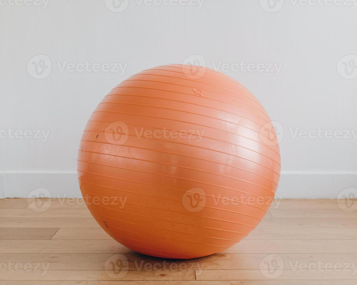 A vibrant orange exercise ball on a wooden floor against a light wall. photo