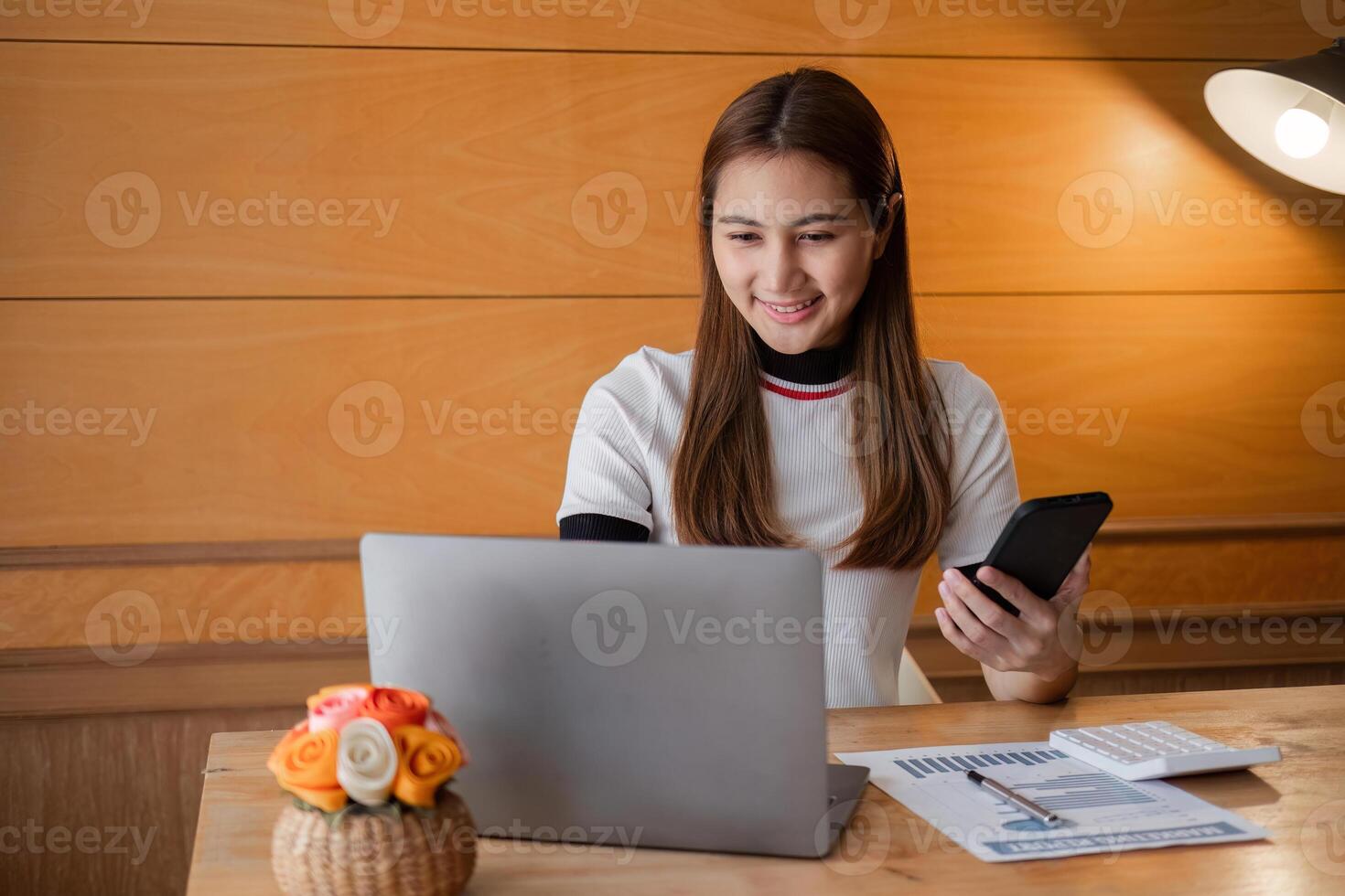 Young Woman Sitting Indoors Focused on Paperwork with Laptop and Smartphone in Modern Home Office Setting photo