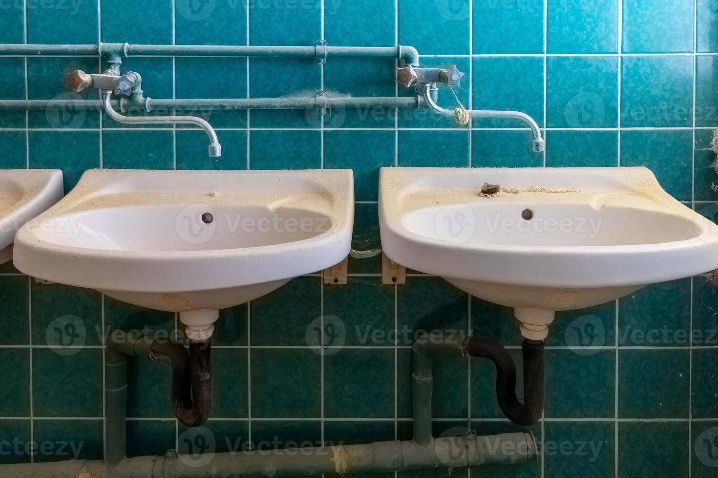 Two white sinks in an outdated bathroom with green tiles showing wear and plumbing details photo