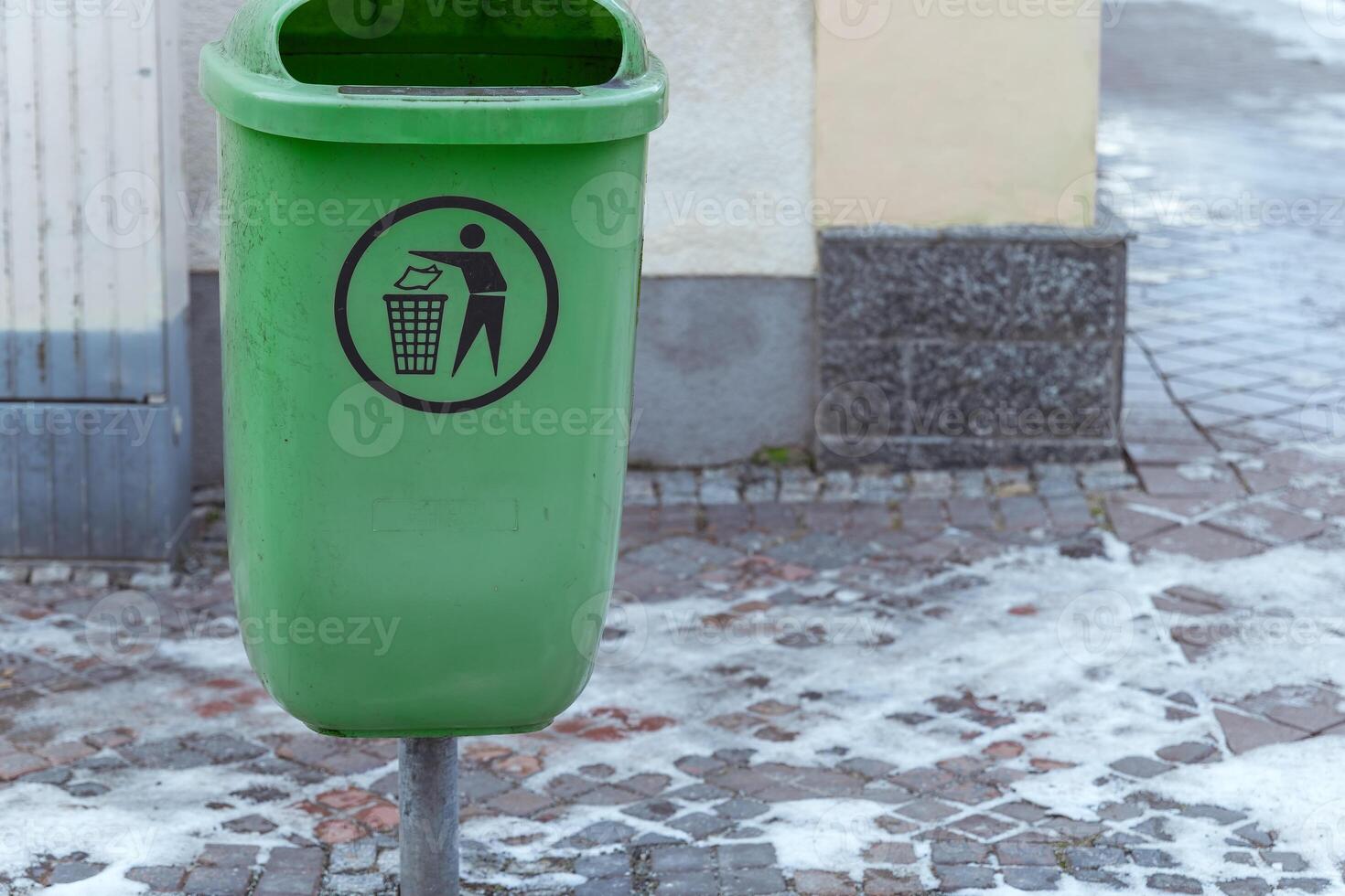 Green trash bin for waste disposal stands on a cobblestone street in winter, promoting cleanliness and recycling efforts photo