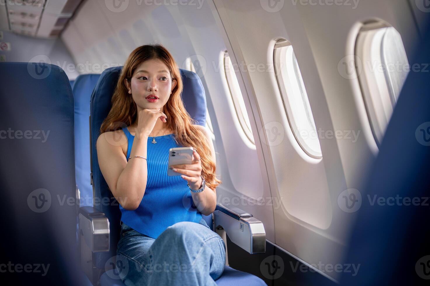 A beautiful Asian woman on a flight, looks out the window at the view while using her smartphone. photo