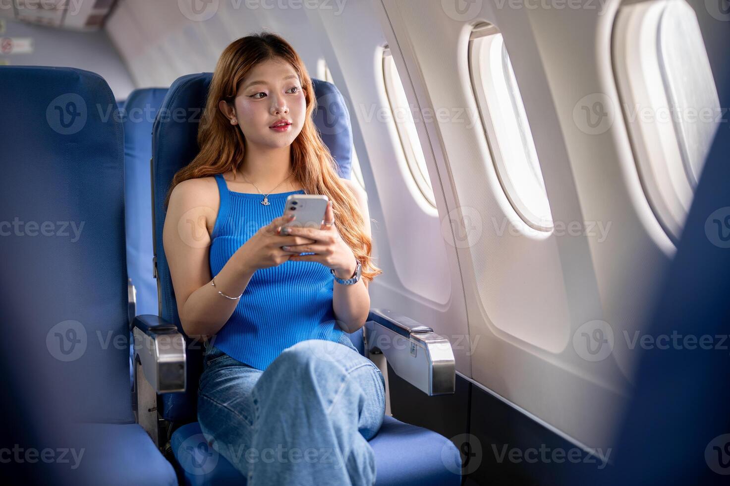A beautiful Asian woman on a flight, looks out the window at the view while using her smartphone. photo
