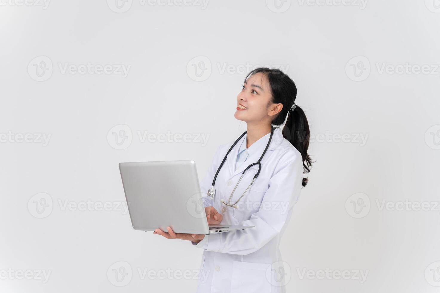 A doctor stands against a white studio background with her laptop, looking upward at an empty space. photo